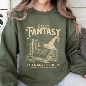 May include: A green sweatshirt with the text "Cozy Fantasy Literary Society Whimsical Tales Bound in Pages" and a graphic of a stack of books with a witch's hat on top.