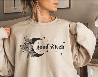 May include: A beige sweatshirt with a black graphic design of a crescent moon, a butterfly, and stars. The text "good witch" is printed on the sweatshirt.
