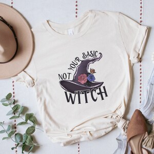 Puede incluir: Una camiseta de color crema con el texto "Not Your Basic Witch" y un sombrero de bruja morado con flores.
