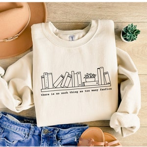 May include: Cream-colored sweatshirt with a black line drawing of a bookshelf and the text "there is no such thing as too many fanfics." The sweatshirt is paired with blue jeans, a tan hat, and sandals.