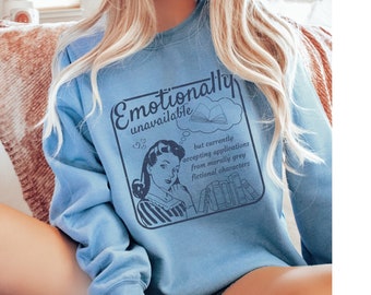May include: Light blue sweatshirt with the text "Emotionally unavailable" and a vintage-style illustration of a woman. The graphic includes the phrase "but currently accepting applications from morally grey fictional characters."