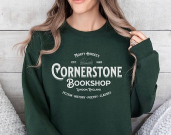 Może przedstawiać: Zielony sweter z białym nadrukiem, na którym widnieje napis "Morty Hindes's Cornerstone Bookshop, Est. 1685, Londyn, Anglia, Fiction - History - Poetry - Classics".