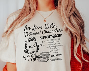 May include: A white t-shirt with a black graphic that says "In Love With Fictional Characters Support Group" and includes a cartoon woman reading a book. The graphic also lists reasons why someone might need to join the support group, such as "Forced Proximity", "Enemies to Lovers", and "Morally Grey Characters".