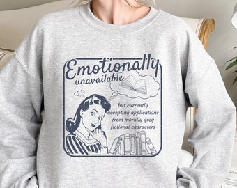 May include: Gray sweatshirt with a vintage-style graphic of a woman reading a book. The text reads "Emotionally unavailable but currently accepting applications from morally grey fictional characters."