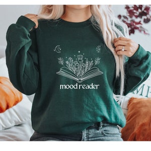 Könnte beinhalten: Ein dunkelgrüner Pullover mit einem weißen Aufdruck eines offenen Buches mit Blumen und Sternen. Der Text "mood reader" ist unter dem Buch gedruckt.