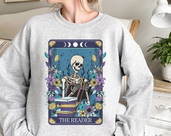 Puede incluir: Una sudadera gris con un estampado gráfico de un esqueleto leyendo un libro. El gráfico está rodeado de flores y tiene el texto "THE READER" en la parte inferior.