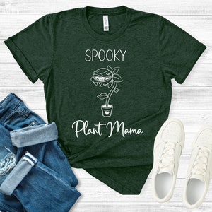 Puede incluir: Una camiseta verde oscuro con un gráfico blanco de una planta con una boca y el texto "Spooky Plant Mama".
