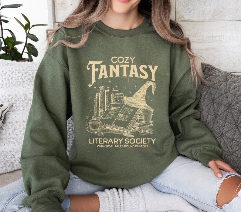 May include: A green sweatshirt with the text "Cozy Fantasy Literary Society Whimsical Tales Bound in Pages" and a graphic of a stack of books with a witch's hat on top.