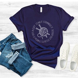 May include: A navy blue t-shirt with a white graphic of a ball of yarn and knitting needles. The text around the graphic reads "It's guts throw it off a cliff stab it strangle it scoop out its guts".