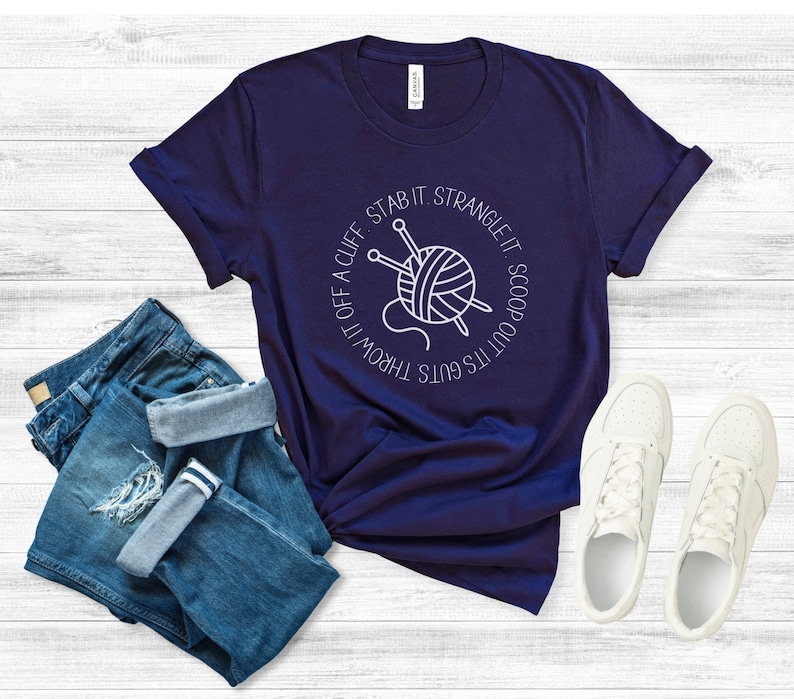 May include: A navy blue t-shirt with a white graphic of a ball of yarn and knitting needles. The text around the graphic reads "It's guts throw it off a cliff stab it strangle it scoop out its guts".