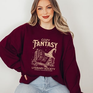 May include: A maroon sweatshirt featuring a graphic design of a stack of books, a witch's hat, and the text "Cozy Fantasy Literary Society Whimsical Tales Bound in Pages".