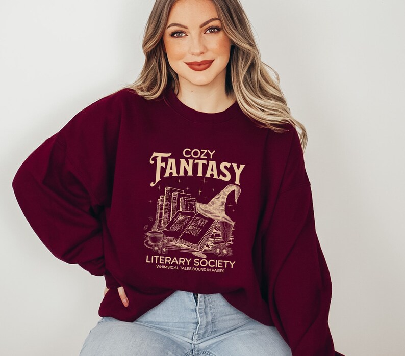 May include: A maroon sweatshirt featuring a graphic design of a stack of books, a witch's hat, and the text "Cozy Fantasy Literary Society Whimsical Tales Bound in Pages".