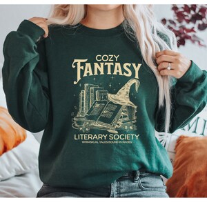 May include: A green sweatshirt with the text "Cozy Fantasy Literary Society Whimsical Tales Bound in Pages" and a graphic of a stack of books, a witch's hat, and stars.