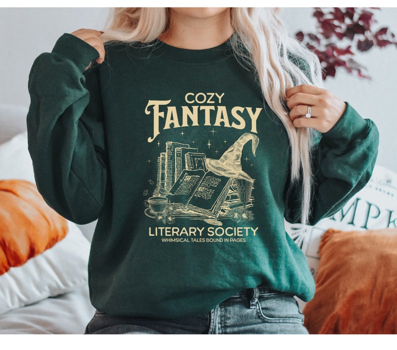 May include: A green sweatshirt with the text "Cozy Fantasy Literary Society Whimsical Tales Bound in Pages" and a graphic of a stack of books, a witch's hat, and stars.