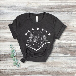May include: A dark gray t-shirt with a white graphic of an open book with mushrooms, trees, and a crescent moon. The book is surrounded by a circle of moons in different phases.