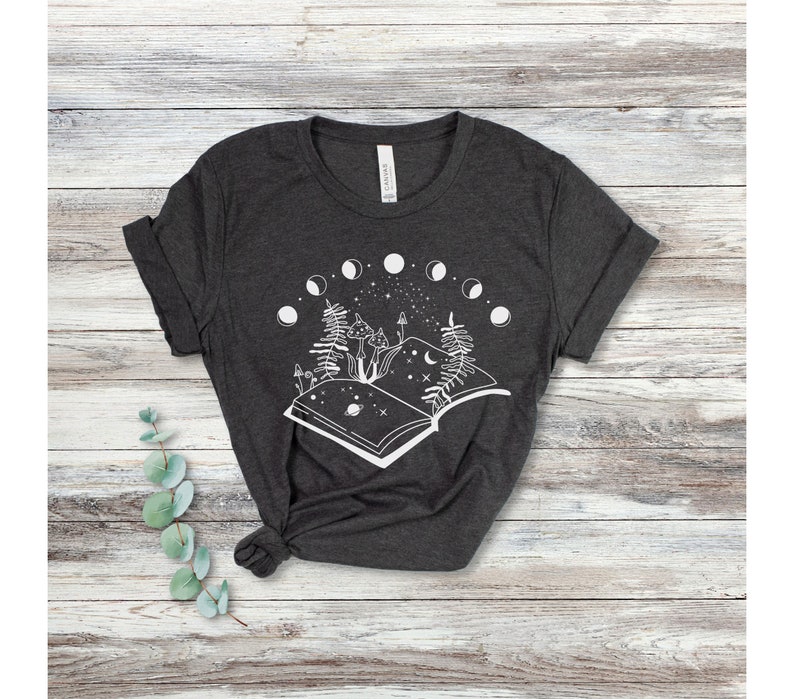 May include: A dark gray t-shirt with a white graphic of an open book with mushrooms, trees, and a crescent moon. The book is surrounded by a circle of moons in different phases.