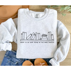 May include: A light gray sweatshirt with a black line drawing of a bookshelf filled with books and a small plant. The text "there is no such thing as too many fanfics" is printed below the bookshelf.