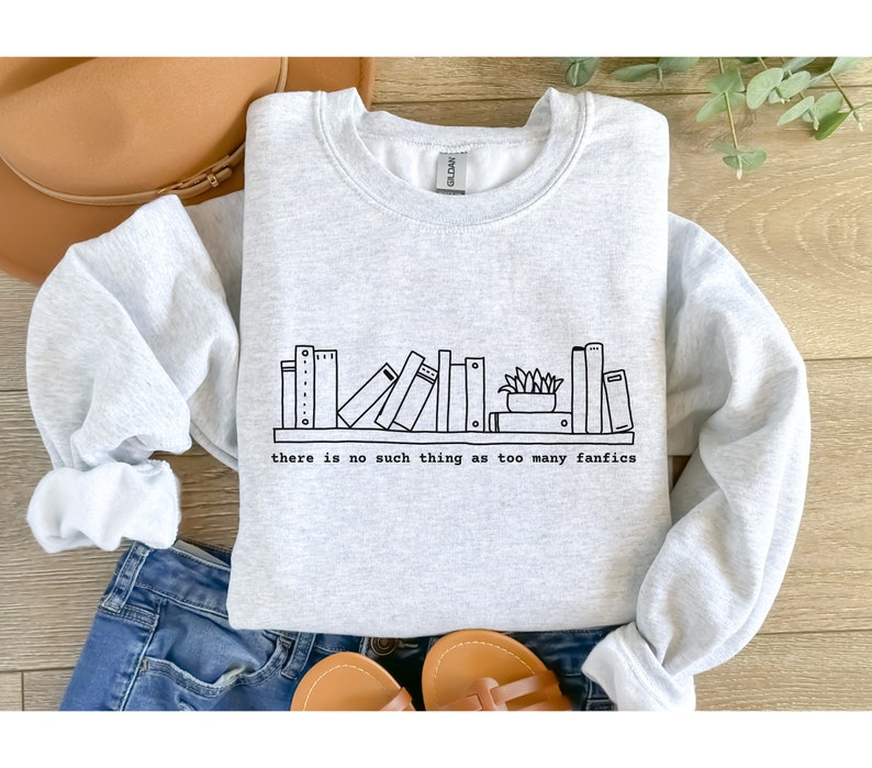 May include: A light gray sweatshirt with a black line drawing of a bookshelf filled with books and a small plant. The text "there is no such thing as too many fanfics" is printed below the bookshelf.
