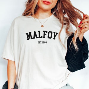 May include: A white t-shirt with black text that reads "MALFOY EST. 1980".