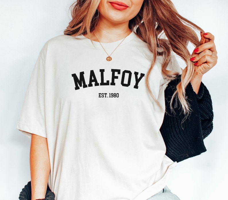 May include: A white t-shirt with black text that reads "MALFOY EST. 1980".