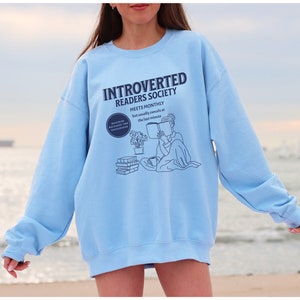 May include: A light blue sweatshirt with the text "Introverted Readers Society Meets Monthly but usually cancels at the last minute" and a line drawing of a person reading a book.