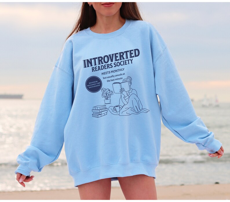 May include: A light blue sweatshirt with the text "Introverted Readers Society Meets Monthly but usually cancels at the last minute" and a line drawing of a person reading a book.