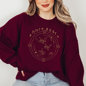 May include: Burgundy sweatshirt with a gold design of origami cranes, stars, and a crescent moon. The design is surrounded by a circle with the text "F M P R B O X Y N" in gold.