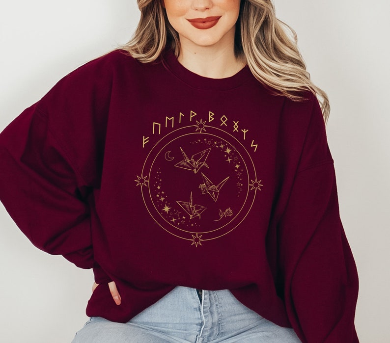 May include: Burgundy sweatshirt with a gold design of origami cranes, stars, and a crescent moon. The design is surrounded by a circle with the text "F M P R B O X Y N" in gold.