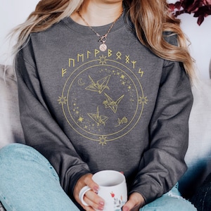 May include: Gray sweatshirt with a gold circle design featuring origami cranes and stars. The text "F M P P R B O X Y S" is written around the circle.