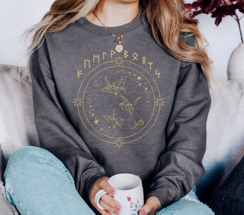 May include: Gray sweatshirt with a gold circle design featuring origami cranes and stars. The text "F M P P R B O X Y S" is written around the circle.