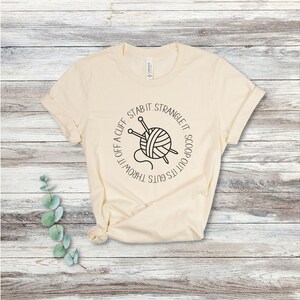 May include: A beige t-shirt with a black and white graphic of a ball of yarn with knitting needles and the text "It's got its guts thrown off a cliff. Stab it. Strangle it. Scoop it out."