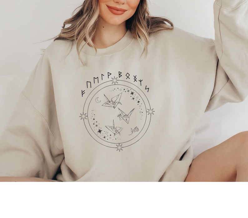 May include: A beige sweatshirt with a black graphic design featuring a circle with stars, a crescent moon, and three origami cranes. The text "F M P P B O X Y S" is written around the circle.