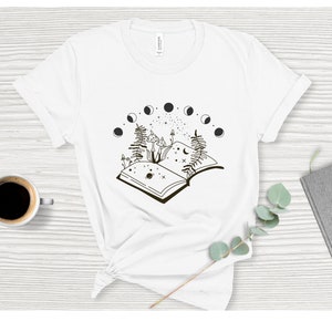 May include: White t-shirt with a black and white illustration of an open book with mushrooms growing from it. The book is surrounded by a crescent moon and stars.