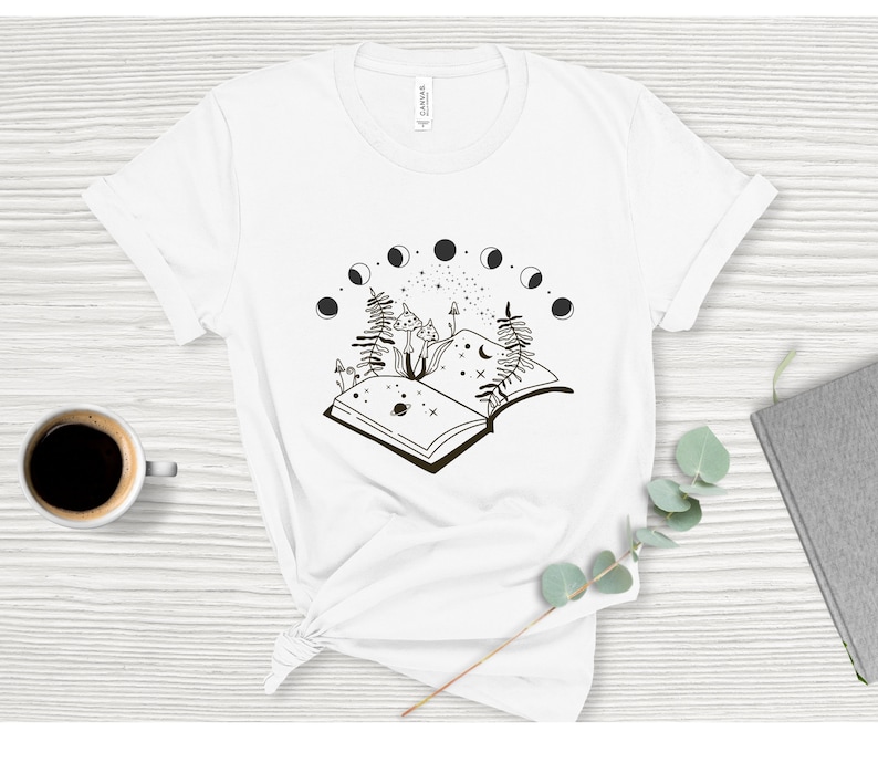 May include: White t-shirt with a black and white illustration of an open book with mushrooms growing from it. The book is surrounded by a crescent moon and stars.