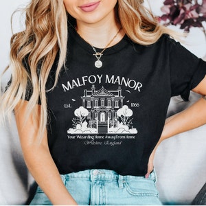 以下が含まれることがあります： マルフォイ・マナーの白いグラフィックがプリントされた黒いTシャツ。大きな家には「マルフォイ・マナー Est. 1066」と書かれた看板と、「Your Wizarding Home Away From Home Wiltshire, England」というテキストが書かれている。