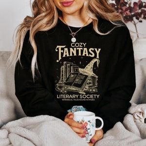 May include: Black sweatshirt with a gold graphic of a stack of books, a witch's hat, and the text "Cozy Fantasy Literary Society Whimsical Tales Bound in Pages".