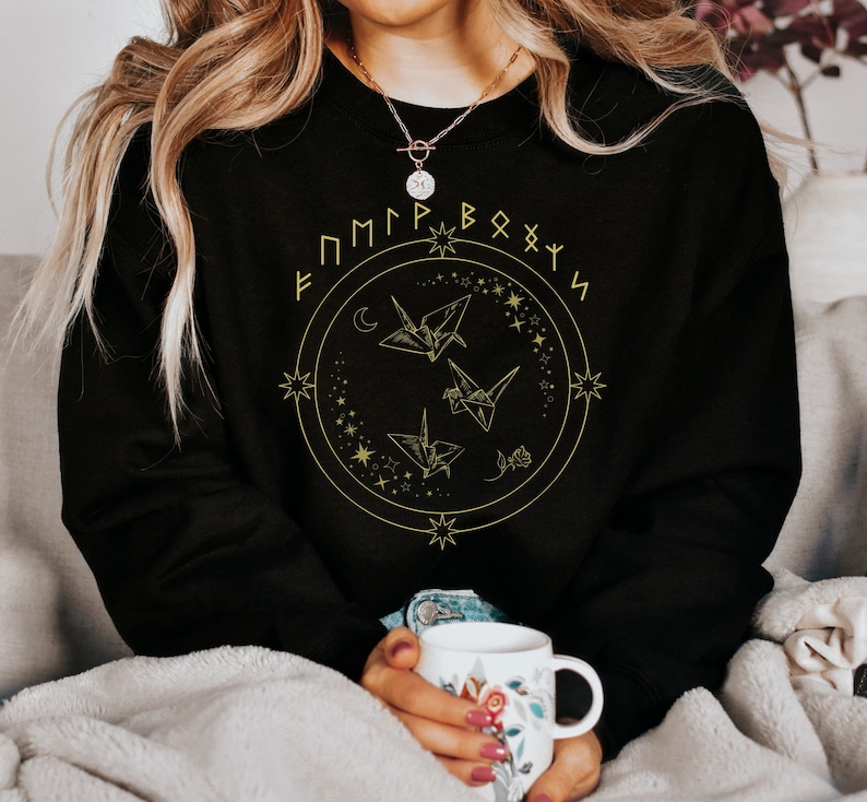 May include: Black sweatshirt with a gold design of origami cranes, stars, and a crescent moon. The design is surrounded by a circle of runes that spell "FMMPRB&Oslash;XYS".