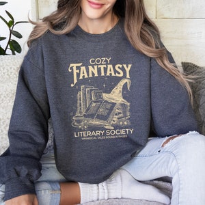 May include: A dark grey sweatshirt with a graphic design of a stack of books, a witch's hat, and the text "Cozy Fantasy Literary Society Whimsical Tales Bound in Pages".