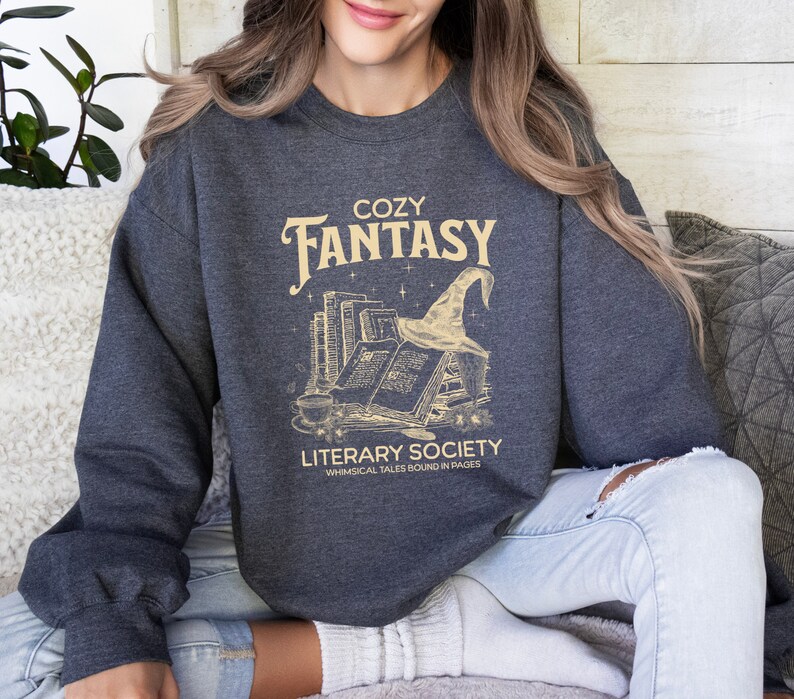 May include: A dark grey sweatshirt with a graphic design of a stack of books, a witch's hat, and the text "Cozy Fantasy Literary Society Whimsical Tales Bound in Pages".