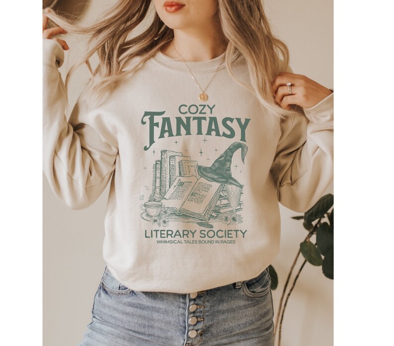 May include: A beige sweatshirt featuring a graphic design of a witch's hat, books, and a cup of tea. The text on the sweatshirt reads "Cozy Fantasy Literary Society Whimsical Tales Bound in Pages".