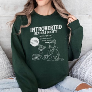 May include: A dark green sweatshirt with the text "Introverted Readers Society" and a graphic of a person reading a book. The text below the graphic says "Meets Monthly but usually cancels at the last minute".