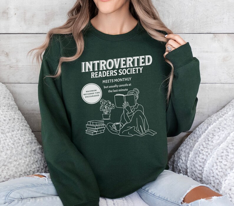 May include: A dark green sweatshirt with the text "Introverted Readers Society" and a graphic of a person reading a book. The text below the graphic says "Meets Monthly but usually cancels at the last minute".