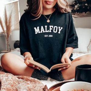 May include: A black oversized t-shirt with white text that reads "MALFOY EST. 1980". The person wearing the shirt is sitting on a bed and reading a book.