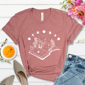 May include: A mauve t-shirt with a white graphic design of an open book with a crescent moon, stars, and two mushrooms. The book is surrounded by a circle of crescent moons.
