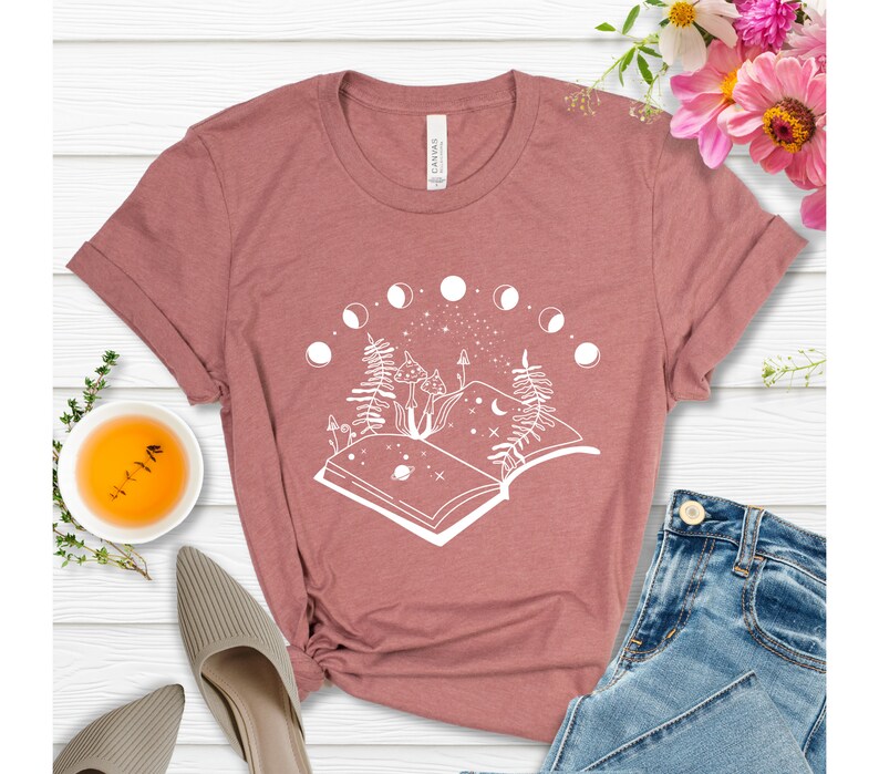 May include: A mauve t-shirt with a white graphic design of an open book with a crescent moon, stars, and two mushrooms. The book is surrounded by a circle of crescent moons.