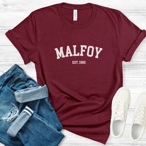 May include: Burgundy t-shirt with white text that reads "MALFOY EST. 1980". The shirt is folded and laid on a white wooden surface with a pair of blue jeans and white trainers.