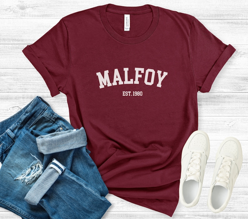 May include: Burgundy t-shirt with white text that reads "MALFOY EST. 1980". The shirt is folded and laid on a white wooden surface with a pair of blue jeans and white trainers.