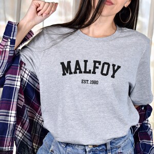 May include: Grey t-shirt with black text that reads "MALFOY EST. 1980".