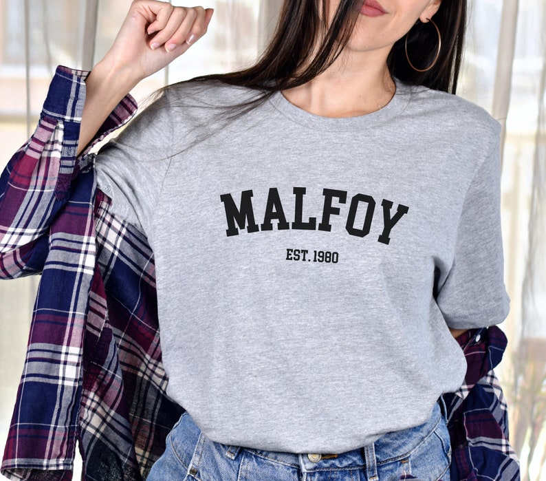 May include: Grey t-shirt with black text that reads "MALFOY EST. 1980".