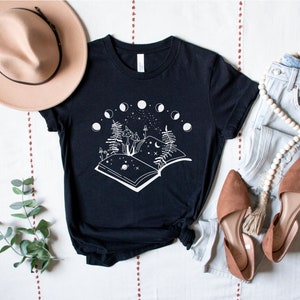 May include: Black t-shirt with a white graphic of an open book with mushrooms, a crescent moon, and stars. The book is surrounded by a circle of eight moons in different phases.